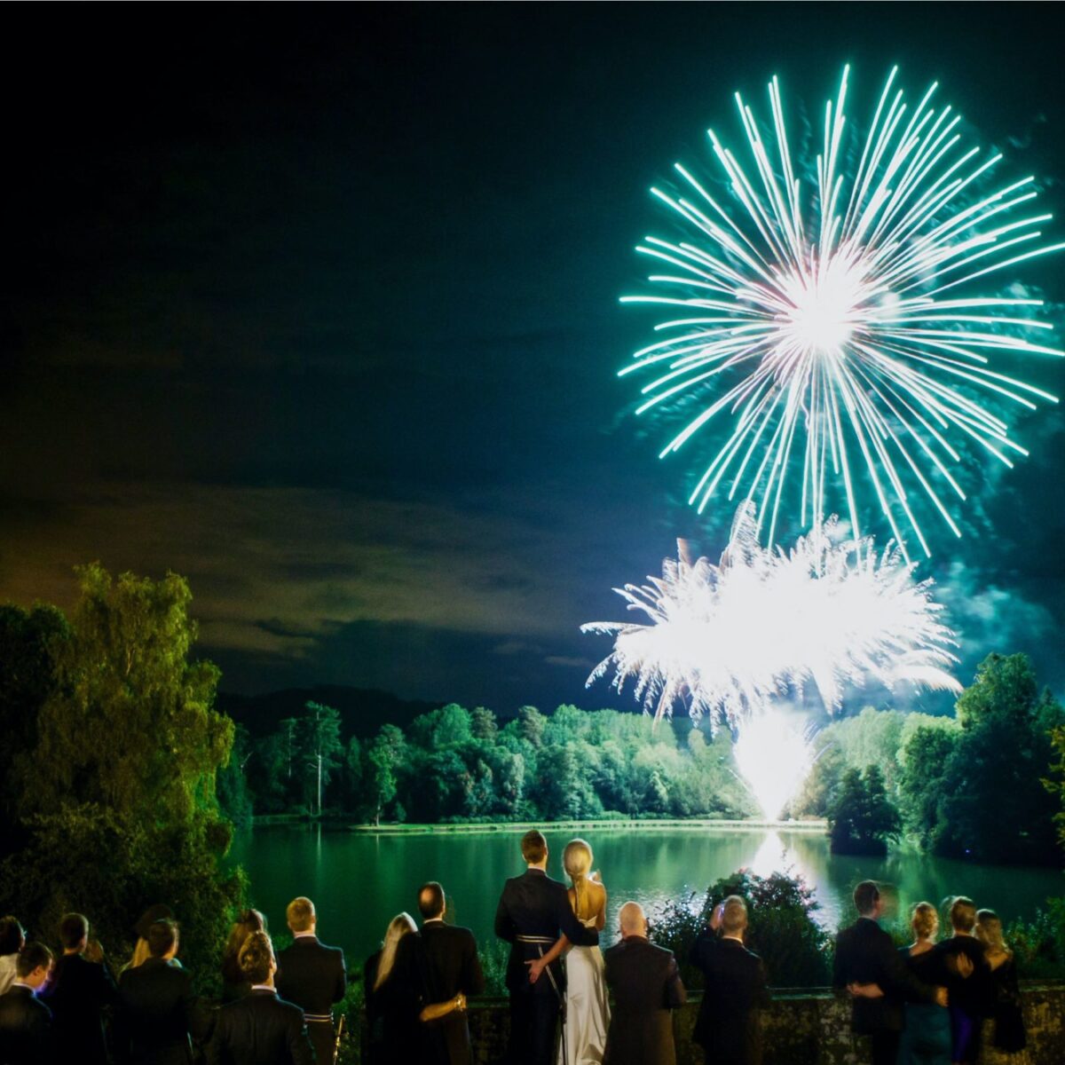 Green and white fireworks over a lake as guests watch